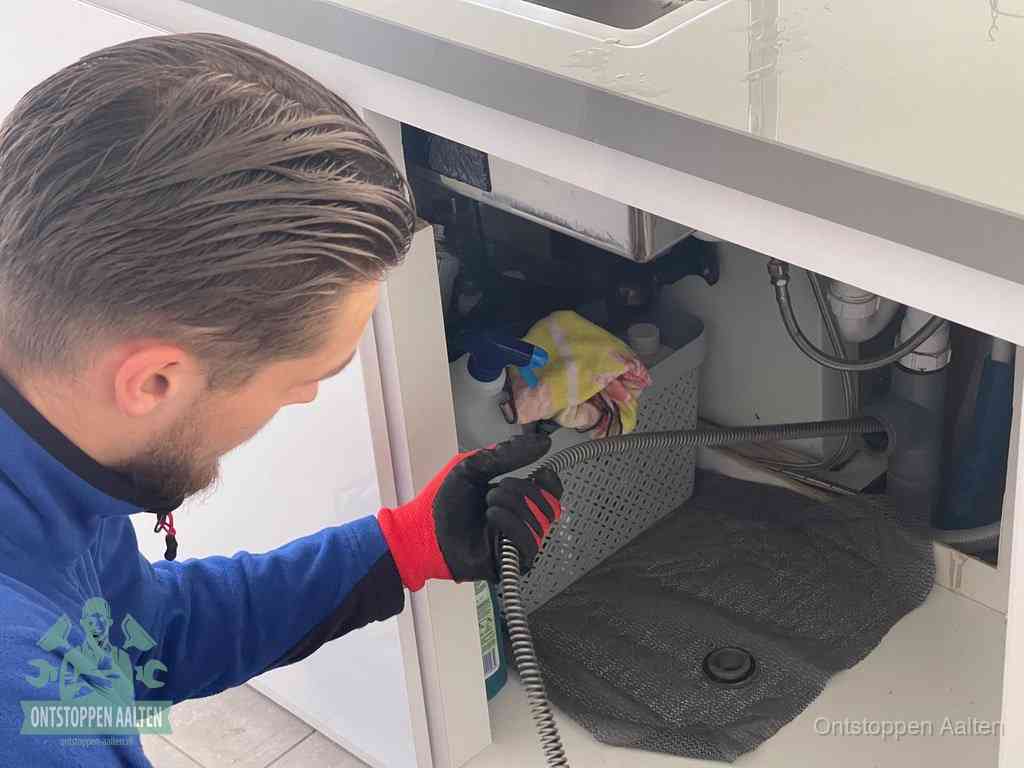 Een monteur voert een flexibele ontstoppingsveer in onder een keukenkastje om een verstopping voorbij de sifon te bereiken in Aalten