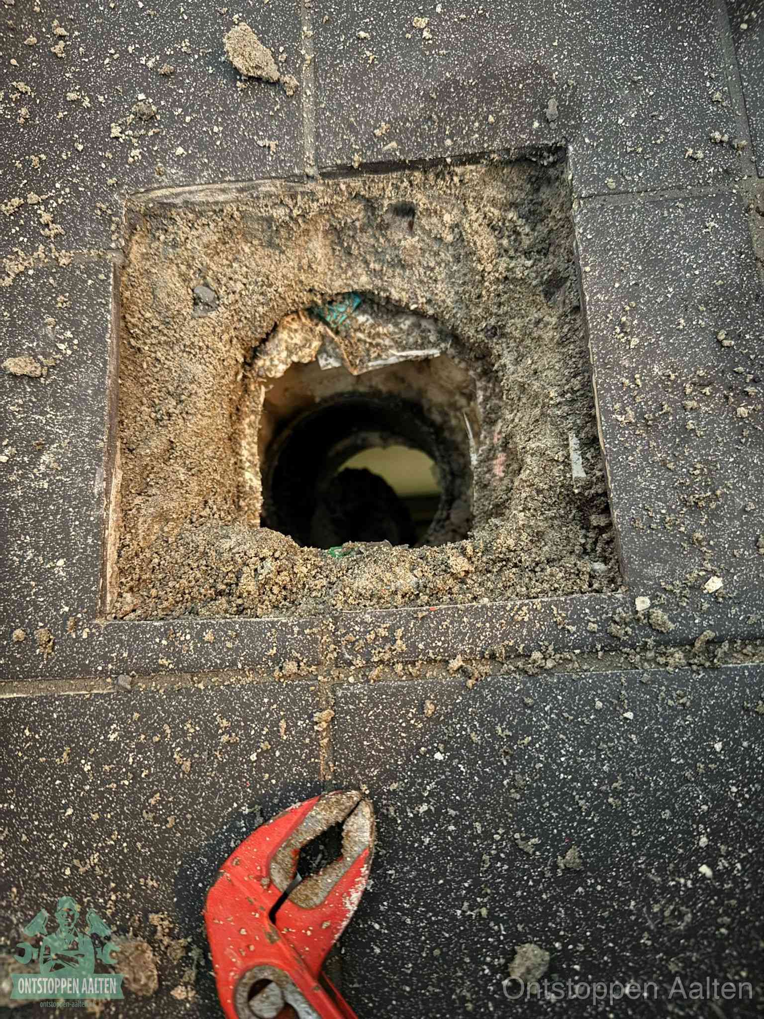 Een geopend rioolputje in een betontegelvloer met losgemaakt rooster en een zichtbare ronde afvoerbuis, het achterhalen waar de afvoerverstopping zit in Aalten