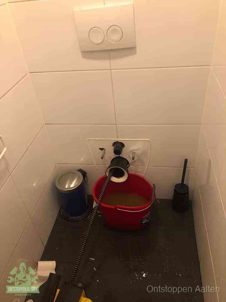 Een ontstopte wand-wc met verwijderde toiletpot waar een ontstoppingsveer in de wanduitgang verdwijnt, het achterhalen waar de verstopping zit in Aalten