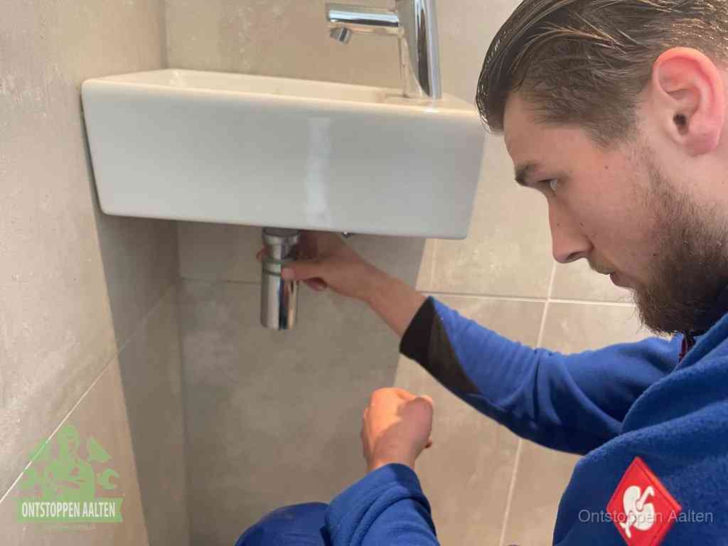 Een monteur draait met de hand de zwanenhalssifon onder een fonteinwasbak in de toiletruimte los om de bron van rioollucht in Aalten te vinden
