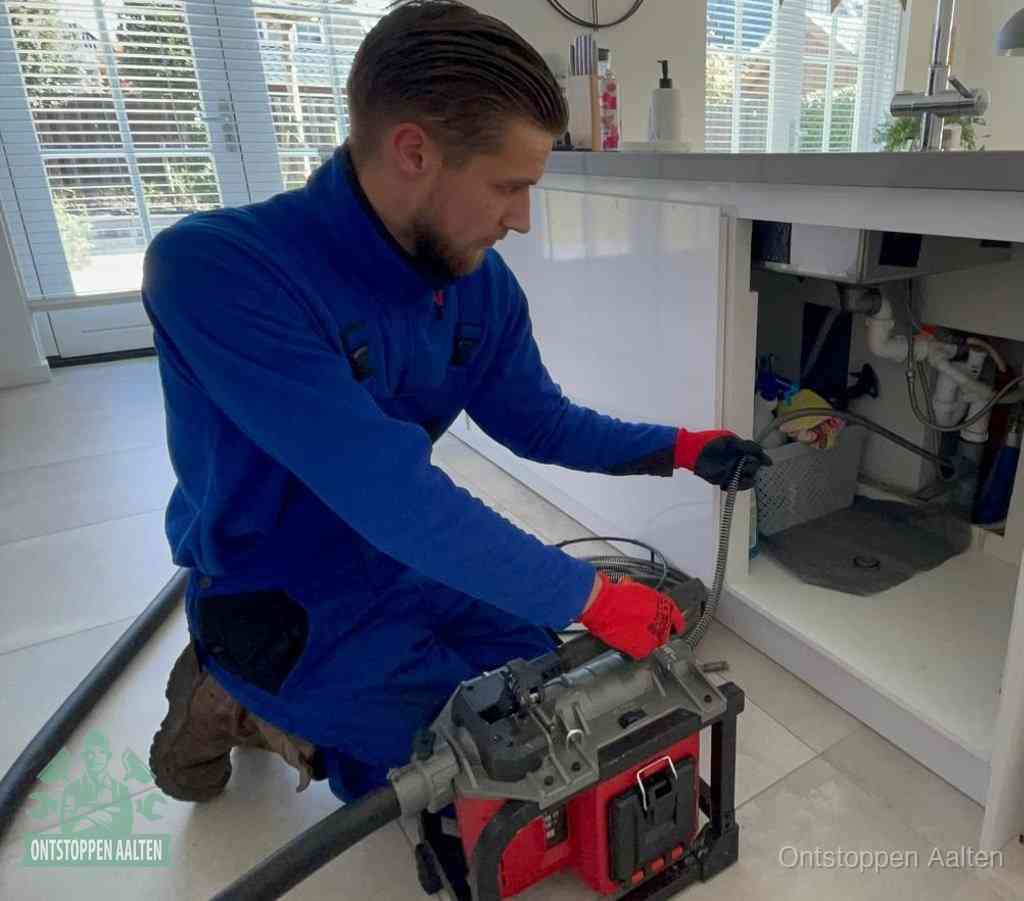 Een monteur met rode handschoenen knielt in een keuken en voert een elektrische ontstoppingsveer in tijdens het professioneel afvoer ontstoppen in Aalten